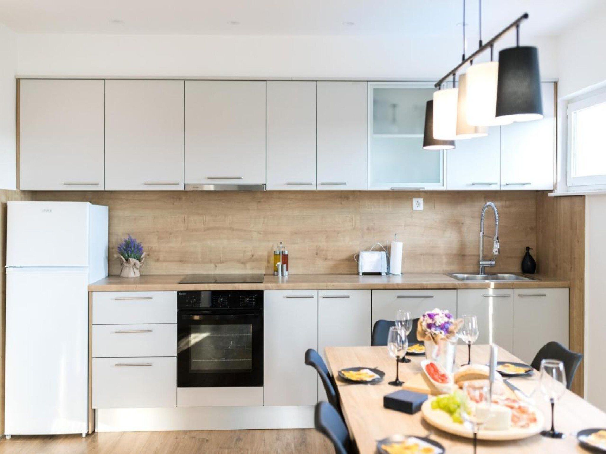 A modern kitchen with white cabinets, a built-in oven, a dishwasher, and a dining area with a wooden table set for a meal.
