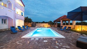 Ein Swimmingpool mit Liegestühlen, einer überdachten Sitzgelegenheit und einem mehrstöckigen Gebäude mit Balkonen.