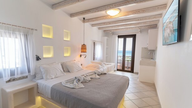 A modern hotel room with a large bed, a small bedside table, a built-in bench, a ceiling light, and a view of the sea through the balcony door. - Nikos Apartments & Studios (Naxos)