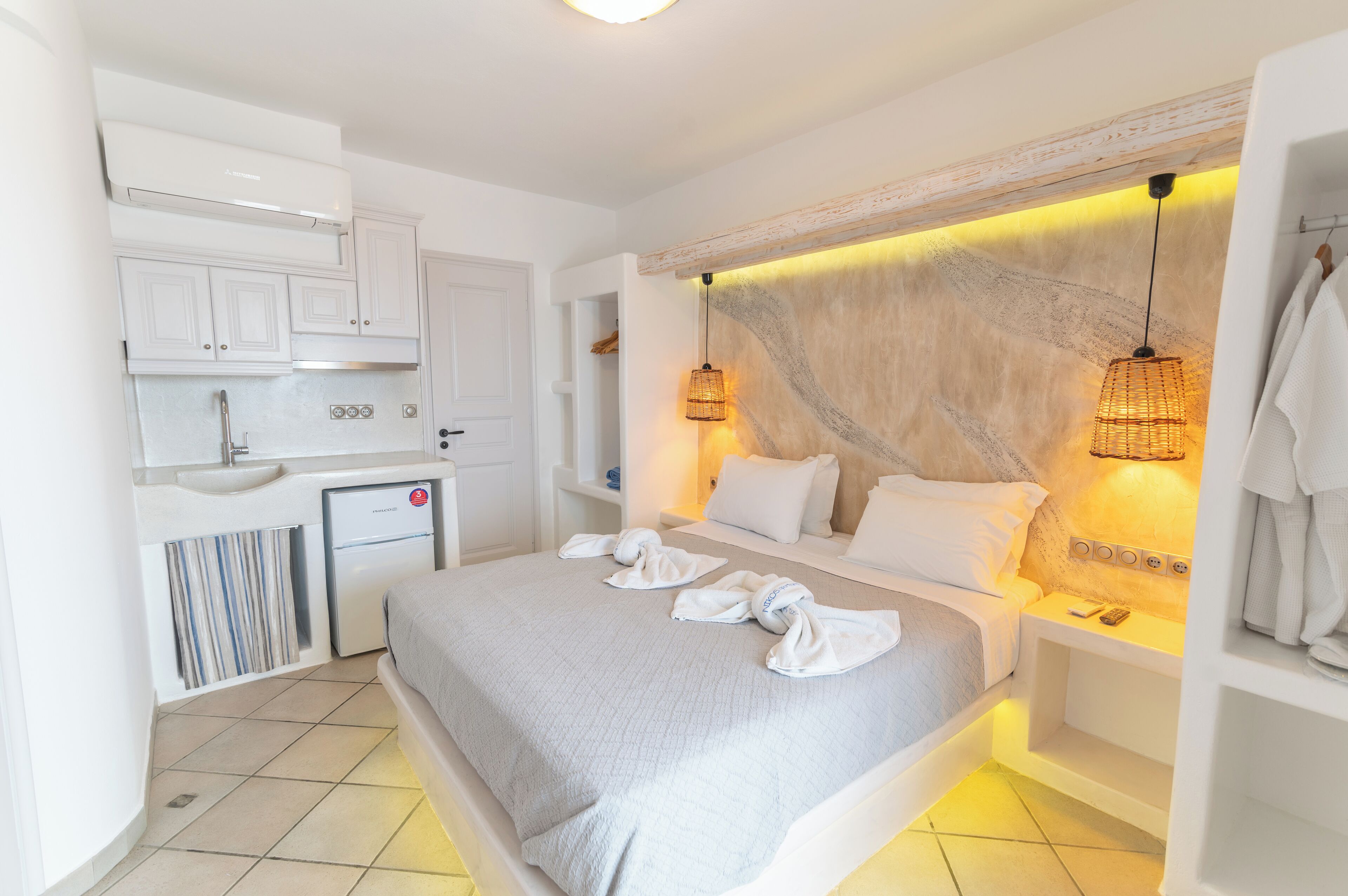 A modern hotel room with a bed, bedside tables, a small kitchenette, and a bathroom.