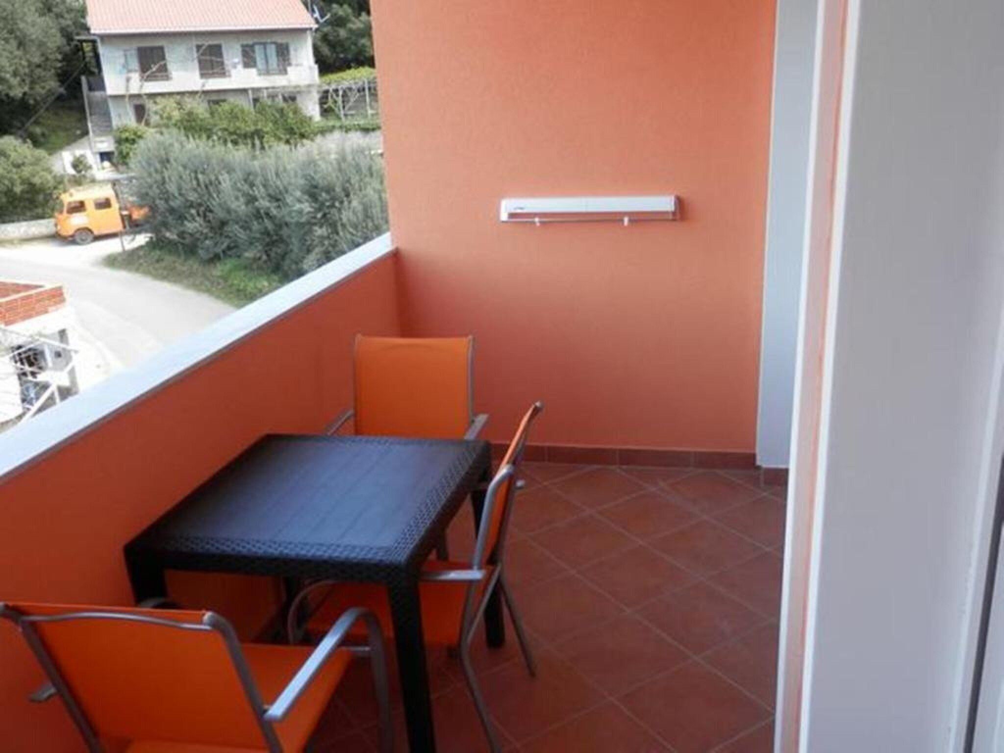A balcony with a table and chairs, a window with a view of a house and a parked car, and a wall-mounted light fixture.