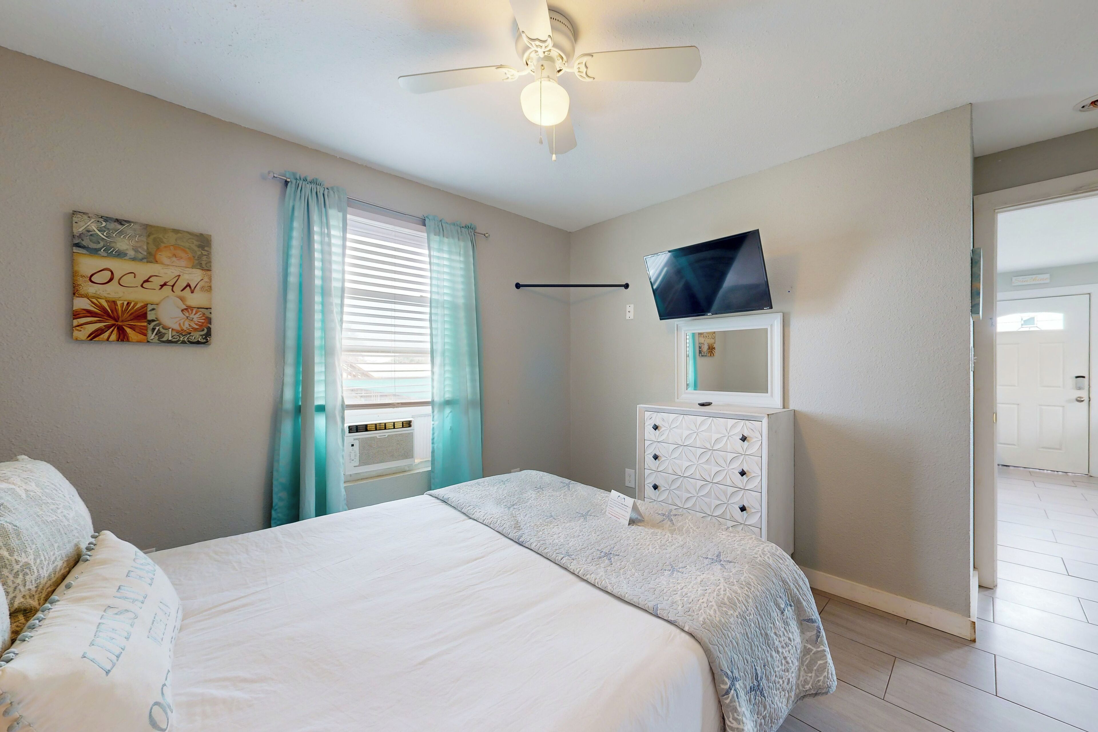 Une chambre à coucher avec un lit, un ventilateur de plafond, un téléviseur posé sur une commode et une fenêtre avec des rideaux.