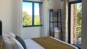 A bedroom with a bed, a bedside lamp, a window with curtains, a ceiling fan, and a view of greenery outside. - Cozy villa between sea and mountains (Figari)