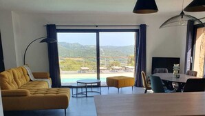 A modern living space with a sofa, a dining table, and bar stools. There is a large window with a view of a pool and hills. - Cozy villa between sea and mountains (Figari)