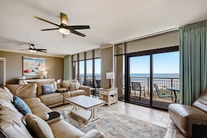 A spacious living room with a large sofa, a coffee table, and a ceiling fan. There is a large window with a view of the ocean.