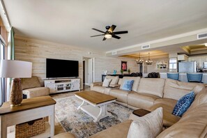 A modern living room with a flat-screen TV, a ceiling fan, and a kitchen in the background.