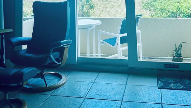 A room with a large window overlooking a beach, a chair, and a small table.
