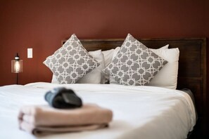 Una cama bien hecha con ropa blanca y dos almohadas estampadas, una lámpara de noche y una toalla doblada sobre la cama.