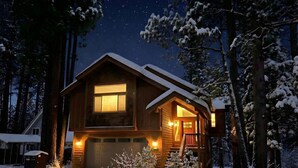 Una cabaña de troncos de dos pisos con garaje, rodeada de árboles cubiertos de nieve y un cielo estrellado.