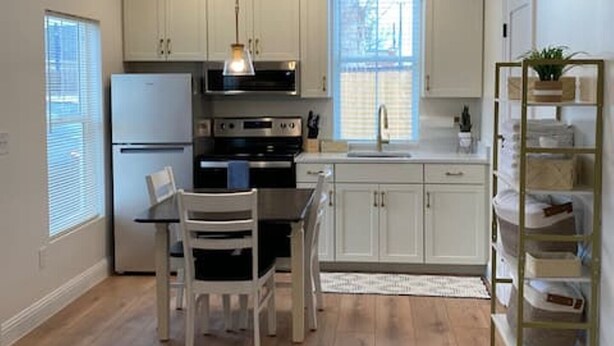A modern kitchen with white cabinets, a stainless steel refrigerator, an oven, a sink, and a dining area with a table and chairs. - Private Urban Cottage in Denver’s RiNo District (Denver)