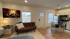 A living room with a brown sofa, a TV mounted on the wall, a dining table with chairs, a kitchen area with cabinets and appliances, and a door with a window. - Private Urban Cottage in Denver’s RiNo District (Denver)