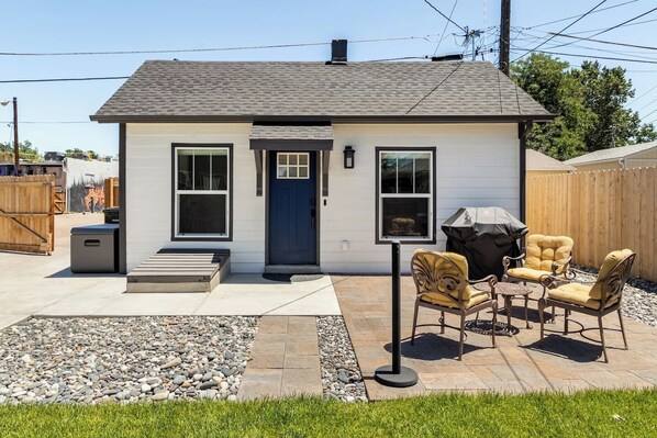 Une maison de plain-pied avec une porte d’entrée, deux fenêtres, un petit porche et un espace extérieur aménagé avec un barbecue.