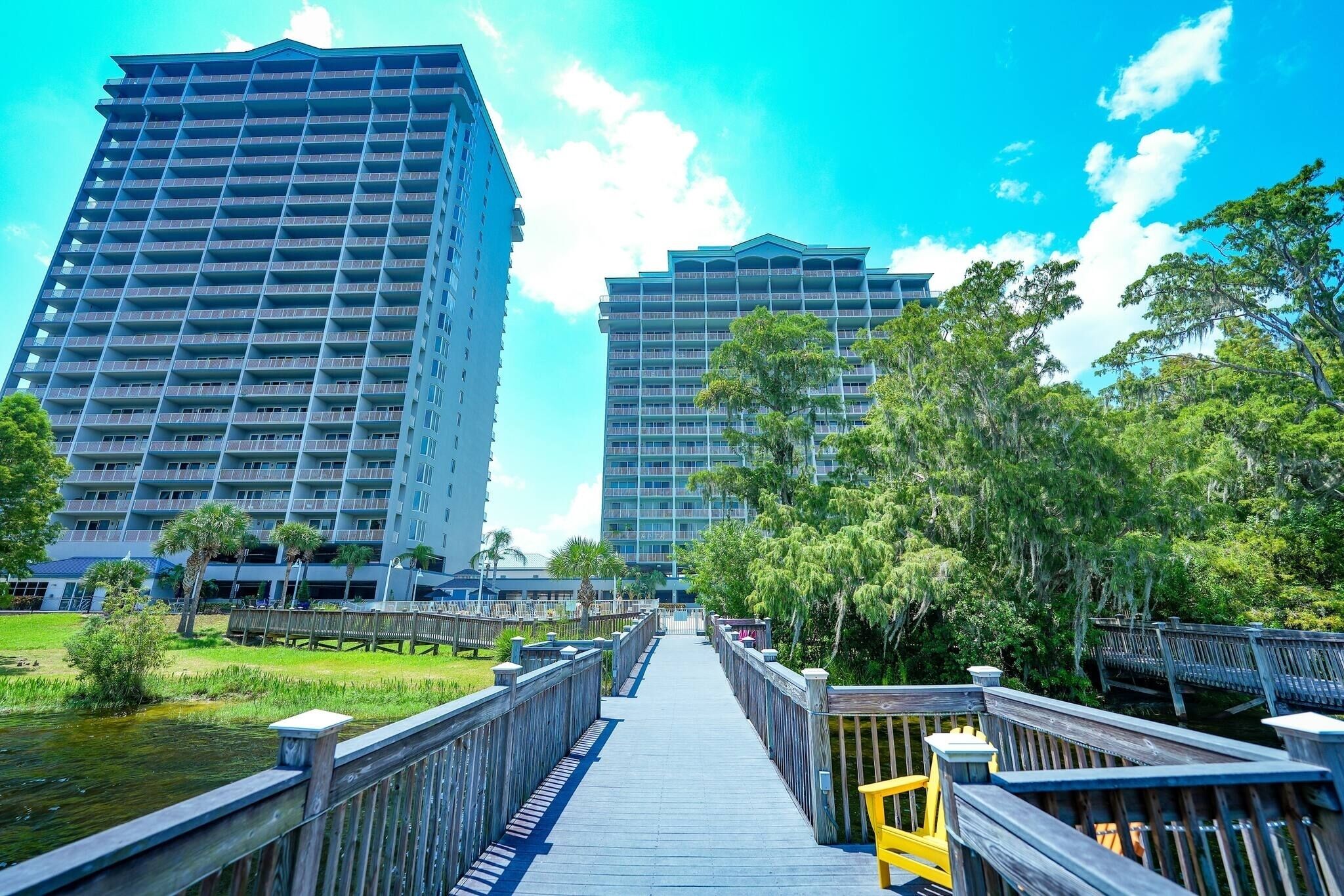 A boardwalk leading to high-rise buildings with balconies, surrounded by greenery and a clear sky.