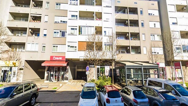 Un immeuble d’appartements à plusieurs étages, doté d’un auvent rouge sur lequel on peut lire l’inscription « PAN-PEK », et d’un espace extérieur abrité avec des sièges.
