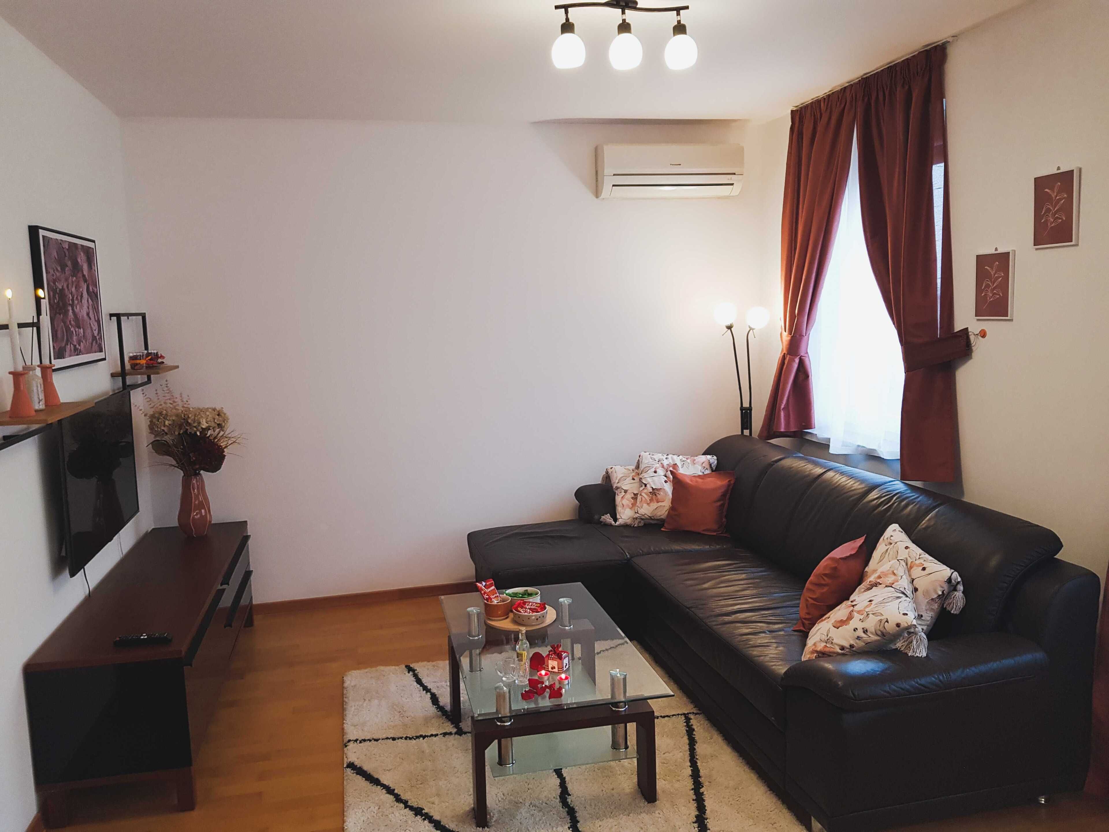 A living room with a black leather sofa, a glass coffee table, a flat-screen TV, a fireplace, and a large window with curtains.