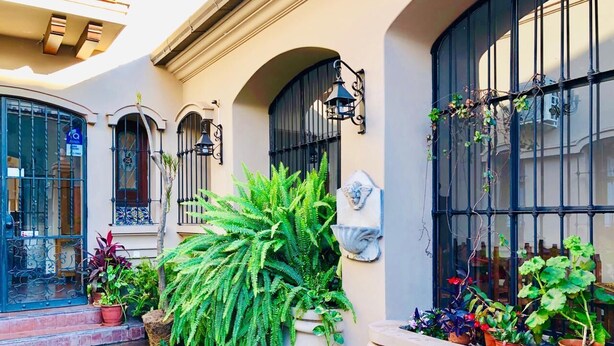 A courtyard with potted plants, a stone floor, and a building with arched windows and black railings. - Posta del Ángel (Salta)