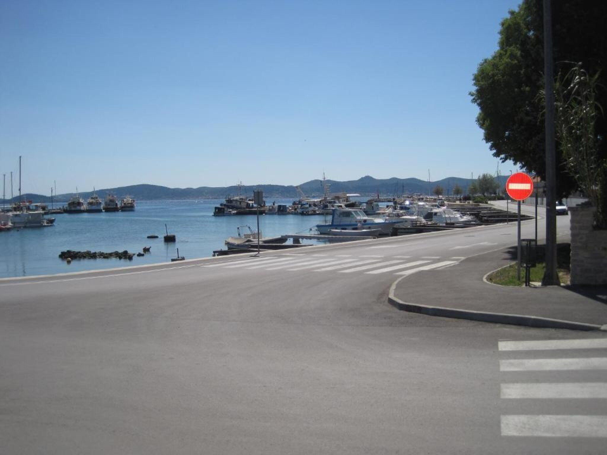 A coastal road with a no entry sign, a pedestrian crossing, and boats docked in the background.