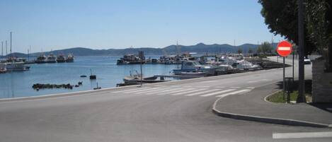 A coastal road with a no entry sign, a pedestrian crossing, and boats docked in the background.