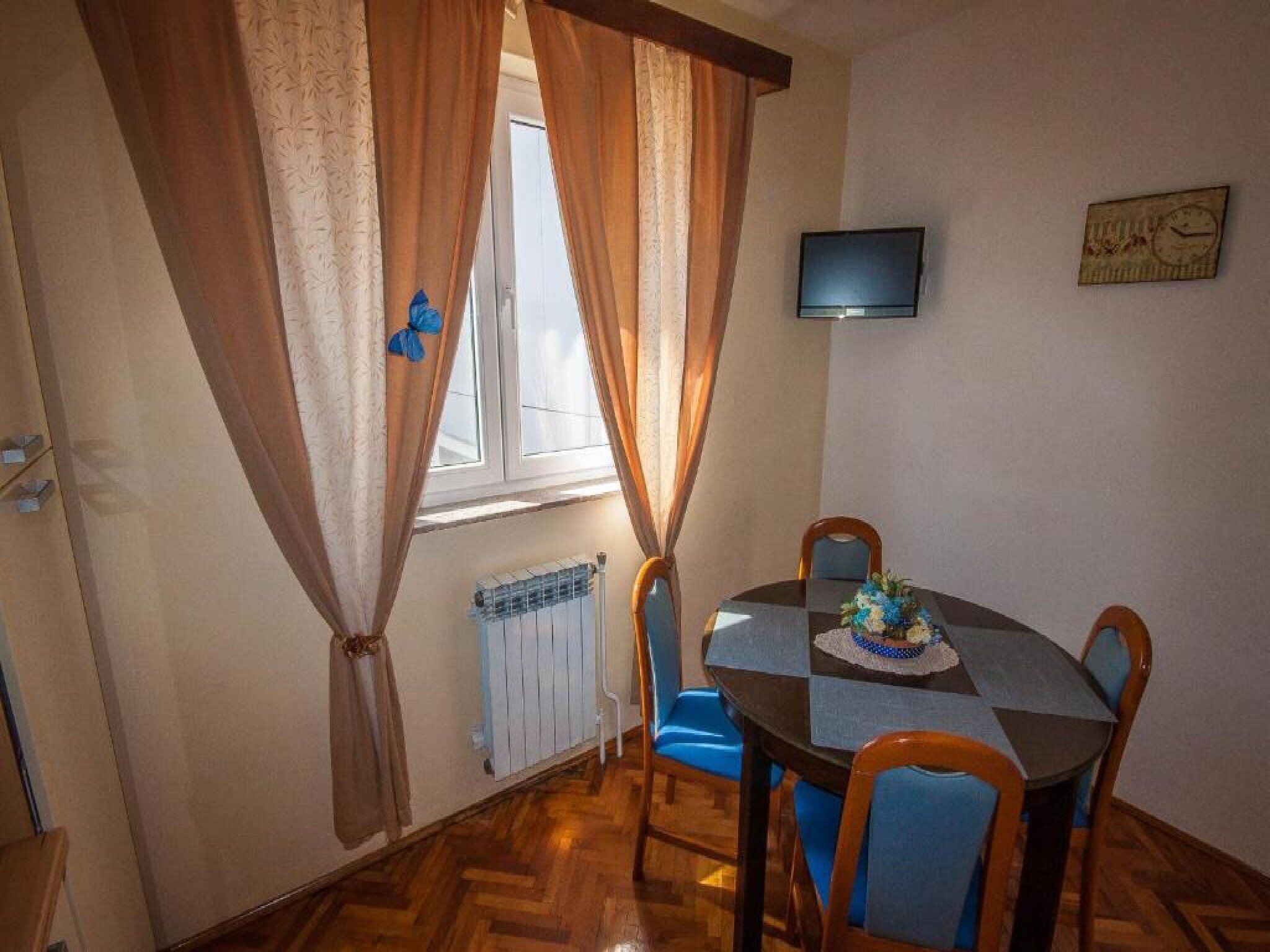 Un coin repas avec une table ronde, des chaises bleues, un téléviseur fixé au mur et une fenêtre avec des rideaux.