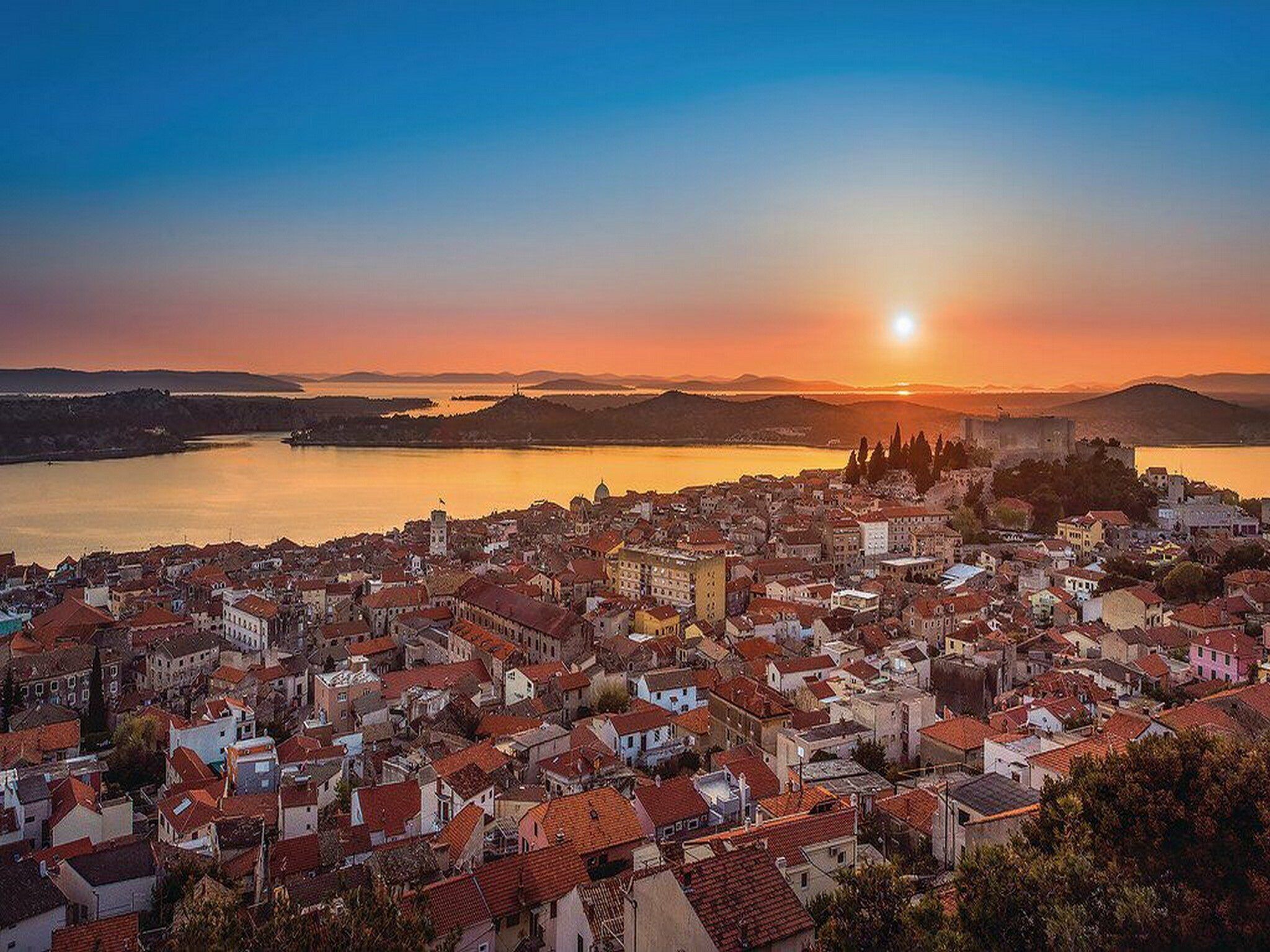 A coastal cityscape during sunset with a prominent castle and dense urban buildings.