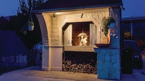 A stone-built outdoor fireplace with a fire burning inside, surrounded by a stone wall and a hanging planter. - Apartment Niko (Sibenik)