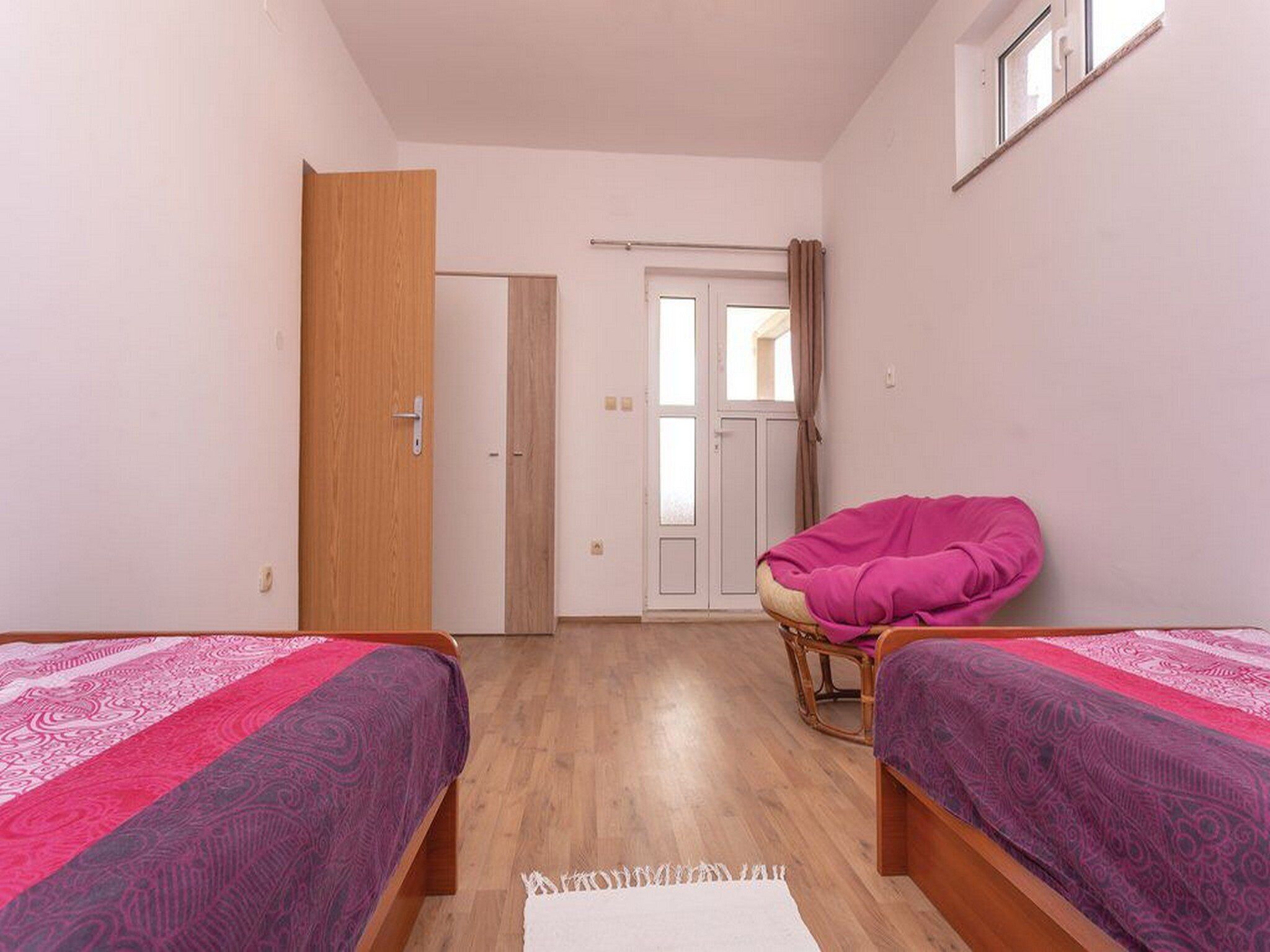 Une chambre avec deux lits, une porte en bois, un mur blanc et une fenêtre avec des rideaux.