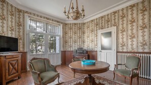 Una habitación con piso de madera, papel tapiz floral, una mesa redonda de comedor con sillas, un televisor y una araña.