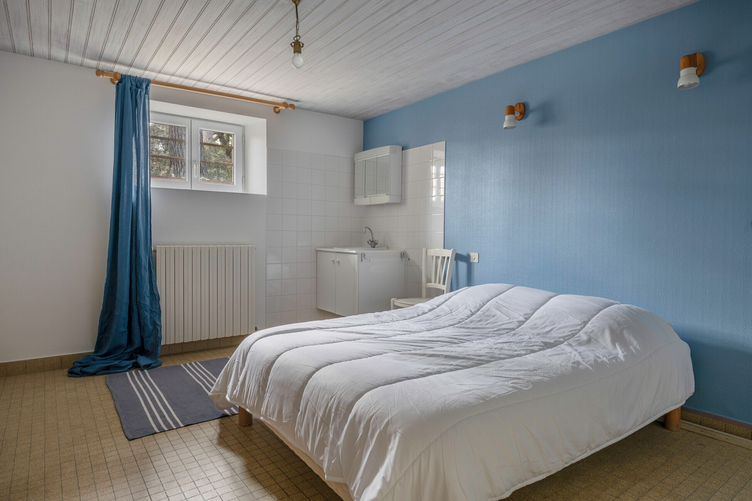 Un dormitorio con una cama grande, una ventana con cortina azul, un radiador blanco, un armario blanco y un espejo en la pared.