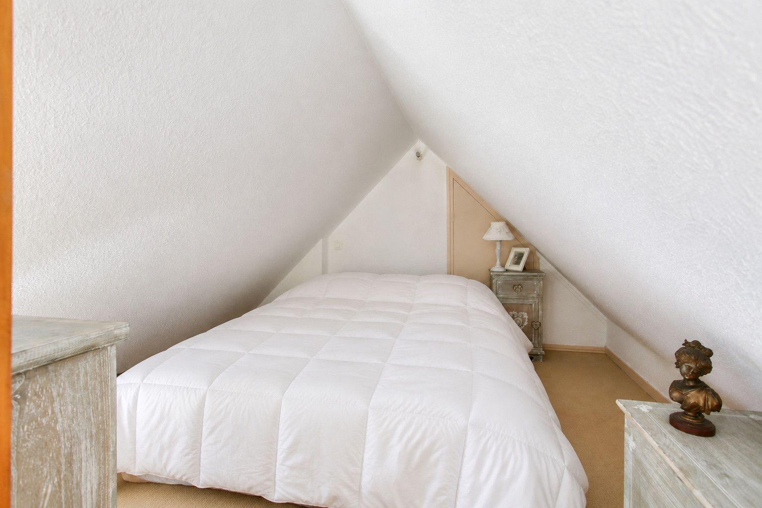 Un dormitorio con una cama blanca, una mesita de noche, una lámpara y un objeto decorativo en un tocador.