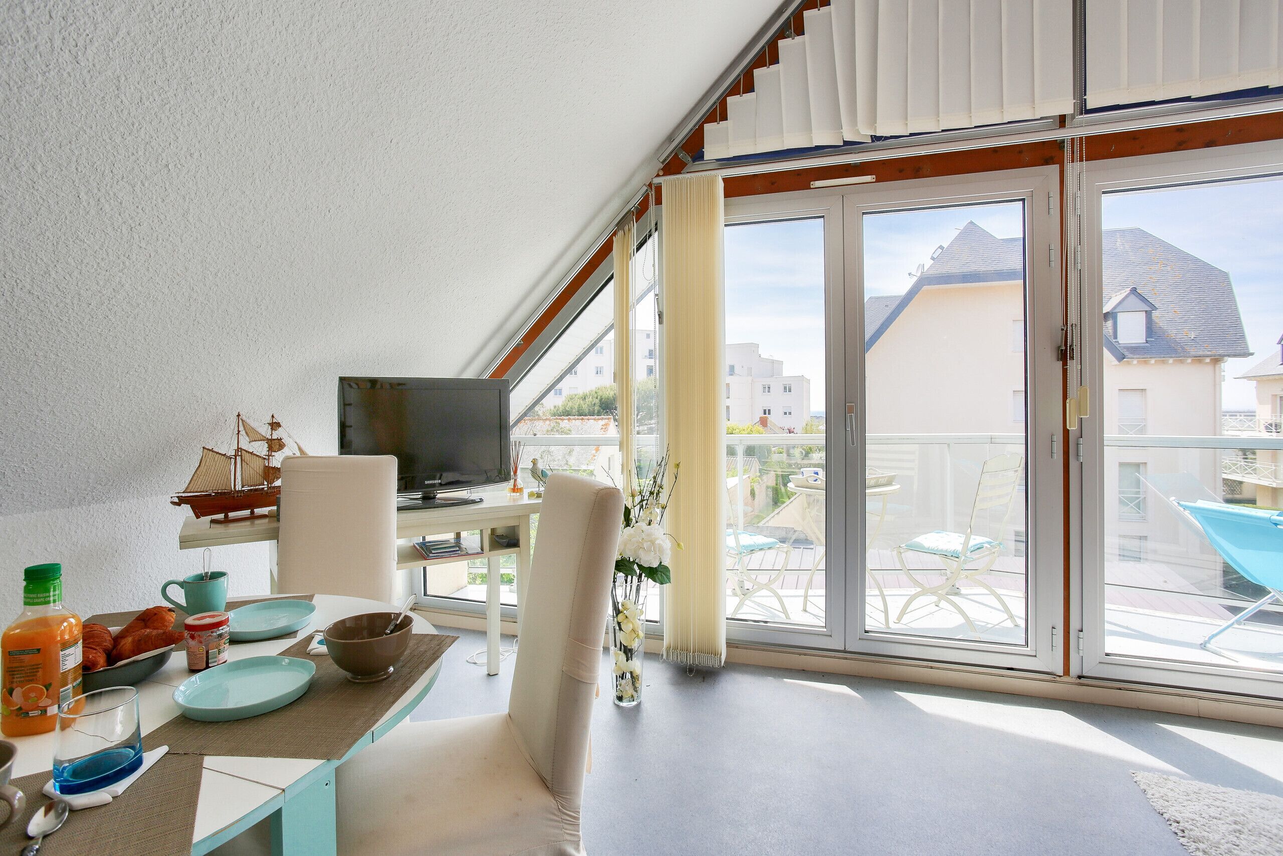A modern room with a balcony, a dining table with breakfast items, a TV, and a view of the neighborhood.