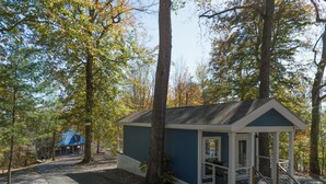 A small cabin with a porch and steps, surrounded by trees and a gravel path.