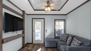 A modern living room with a gray sectional sofa, a flat-screen TV mounted on a wooden shelf, a ceiling fan, and hardwood flooring.