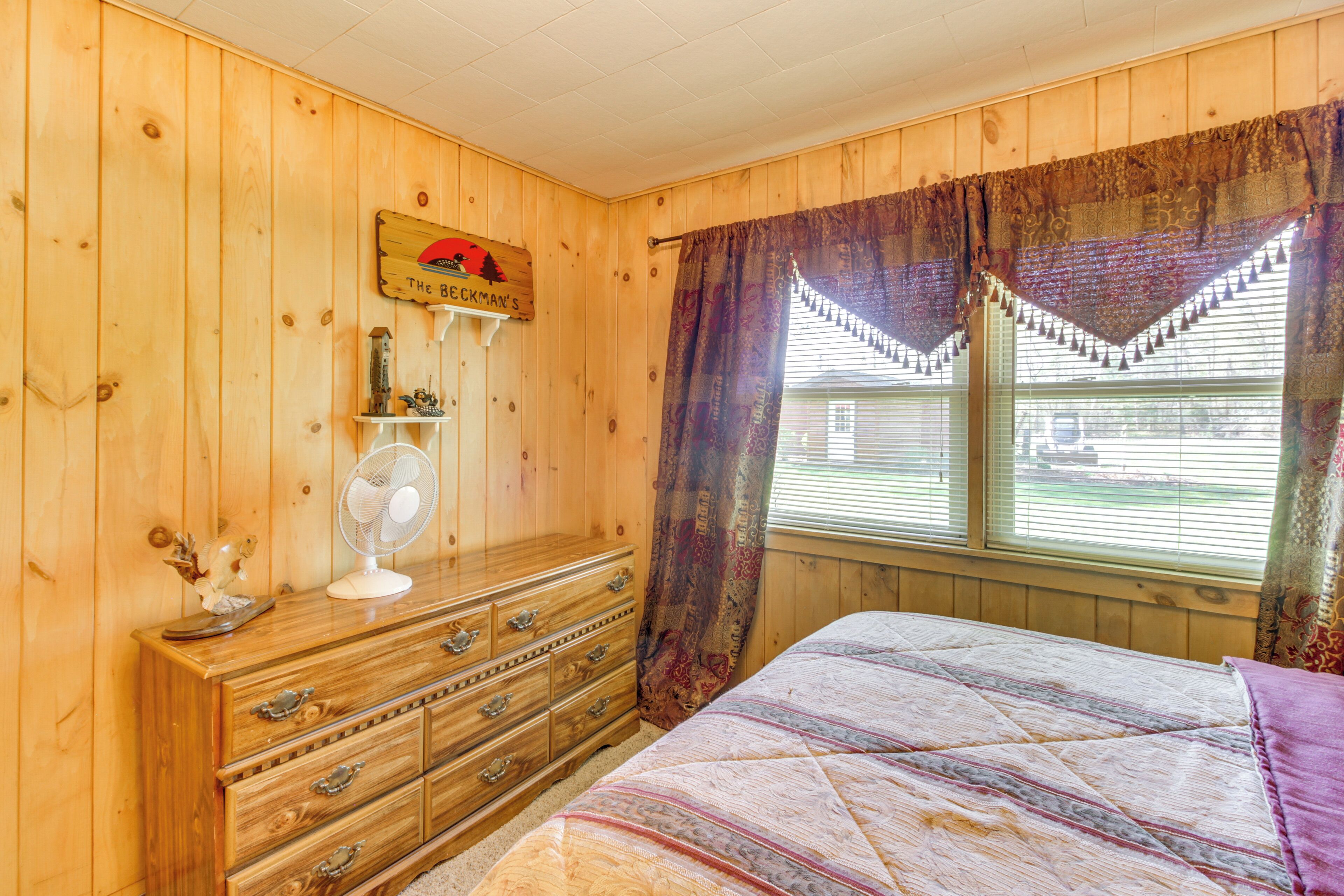 A room with wooden walls, a bed with a patterned bedspread, a dresser with drawers, a fan, and a window with curtains.