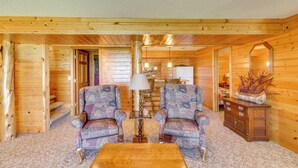 A log cabin interior with two patterned armchairs, a wooden coffee table, a lamp, and a dresser with a mirror. - Private Lakefront Emily Home w/ Yard & Fire Pit! (Emily)