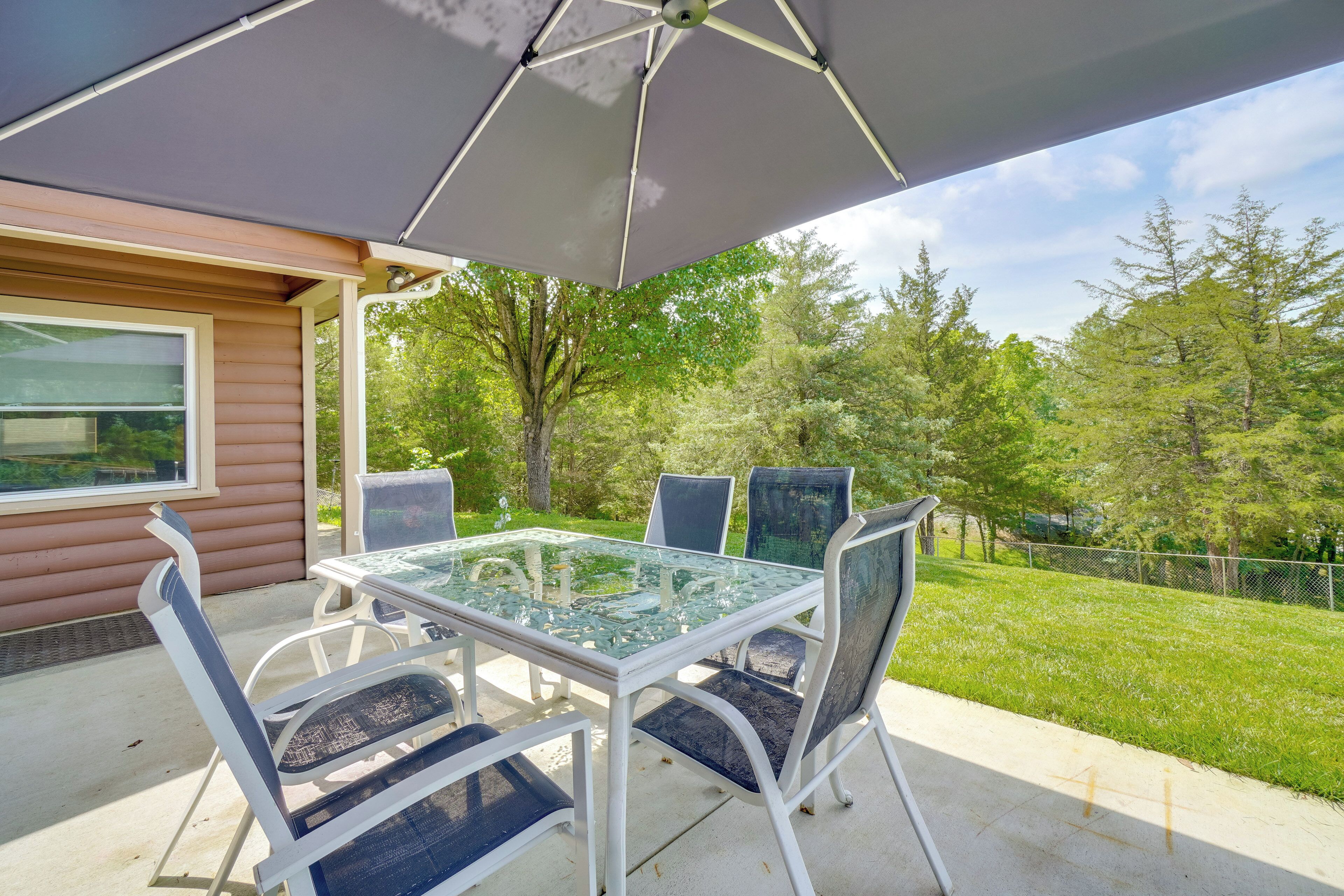 An outdoor patio with a glass table and chairs, a large umbrella, and a view of a grassy area with trees.