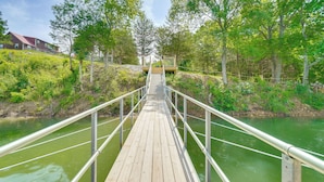 Eine Holzbrücke mit Metallgeländern, die zu einer Plattform mit Geländer führt, umgeben von Grünpflanzen und Bäumen.