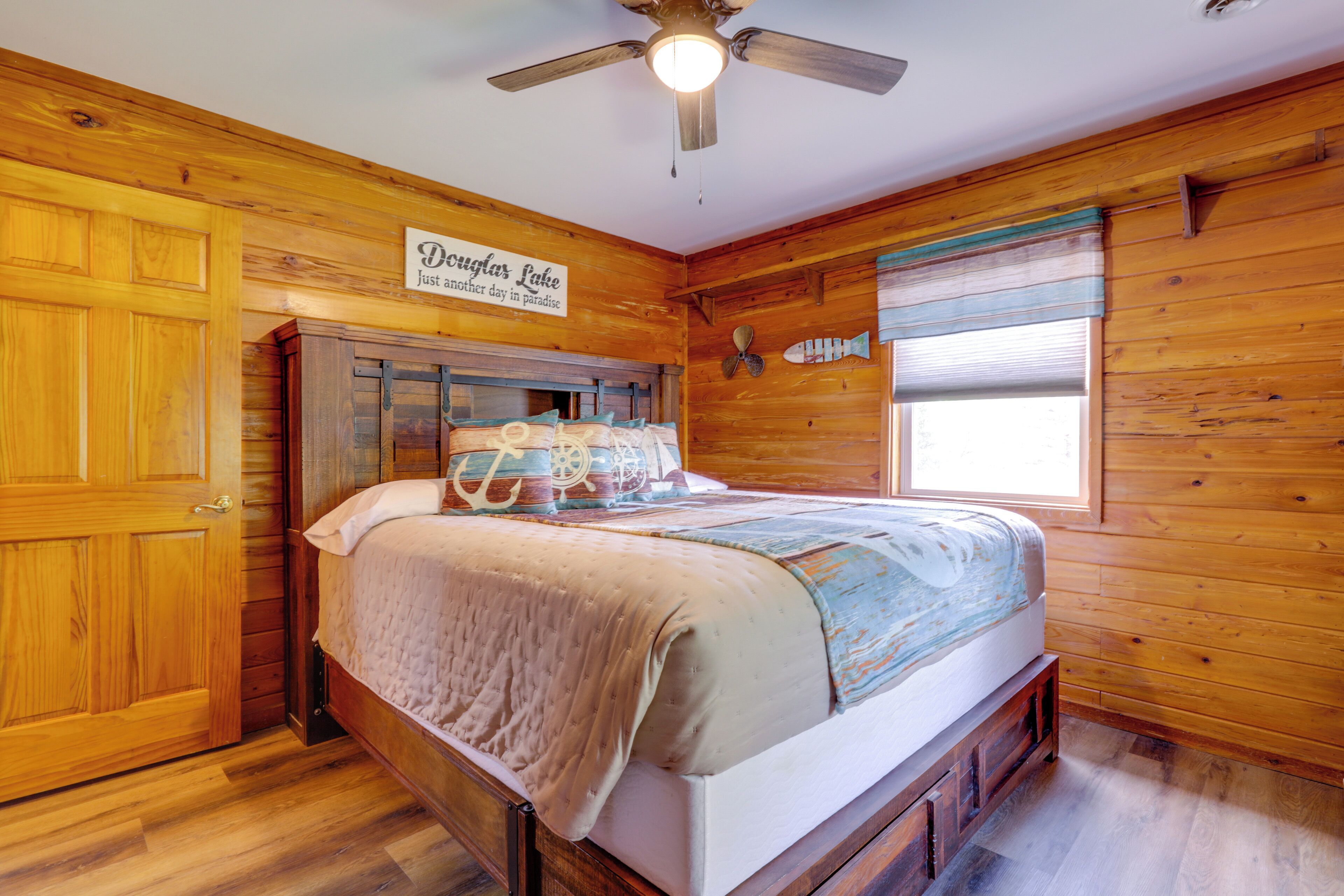 A wooden-paneled room with a bed, a ceiling fan, a window with blinds, and a sign on the wall.