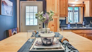 Een eetgedeelte met een houten tafel gedekt voor twee personen, een vaas met bloemen en een keuken op de achtergrond.