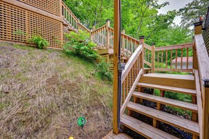 Una terraza de madera con una escalera que sube a un nivel superior, rodeada de vegetación y una casa al fondo.