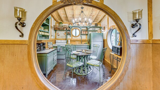 A round mirror with a wooden frame reflecting a kitchen area with green cabinets, a dining table with chairs, and a chandelier. - Magical 2BR Hobbit Themed House Near Gatlinburg (Cosby)