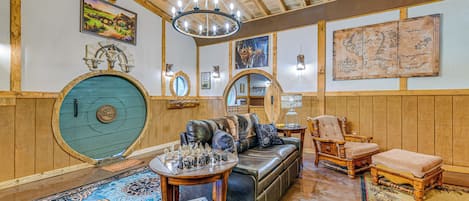 Une pièce chaleureuse avec un plafond en bois, une porte bleue et ronde, un canapé en cuir, une table en bois sur laquelle sont disposés des jeux d’échecs, et une carte accrochée au mur.