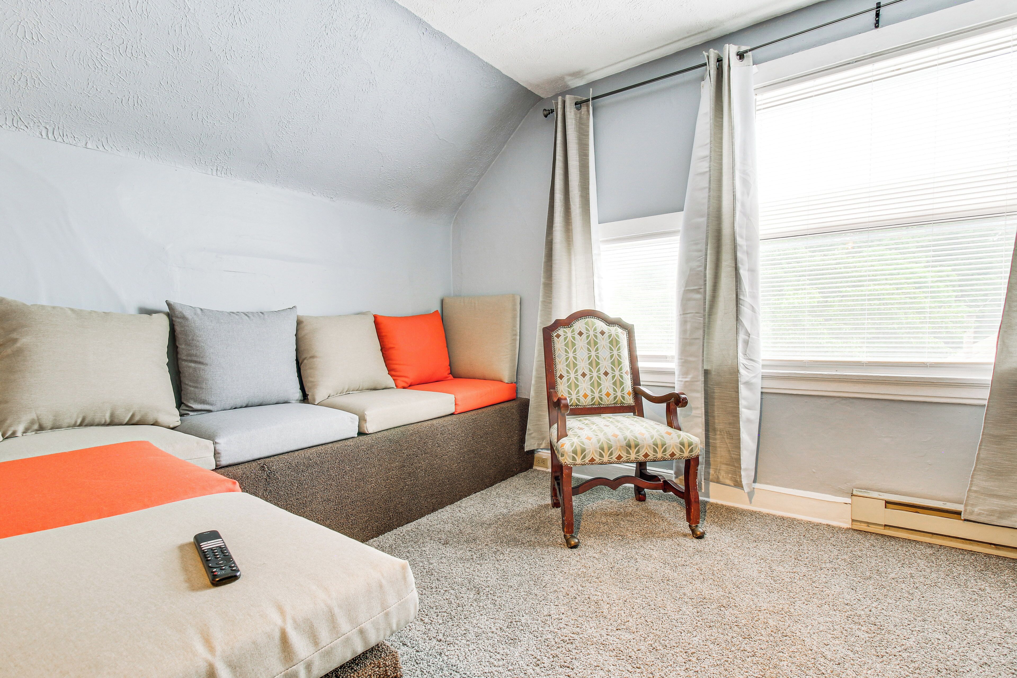 A bedroom with a sofa bed, a wooden chair, a remote control, and a window with curtains.