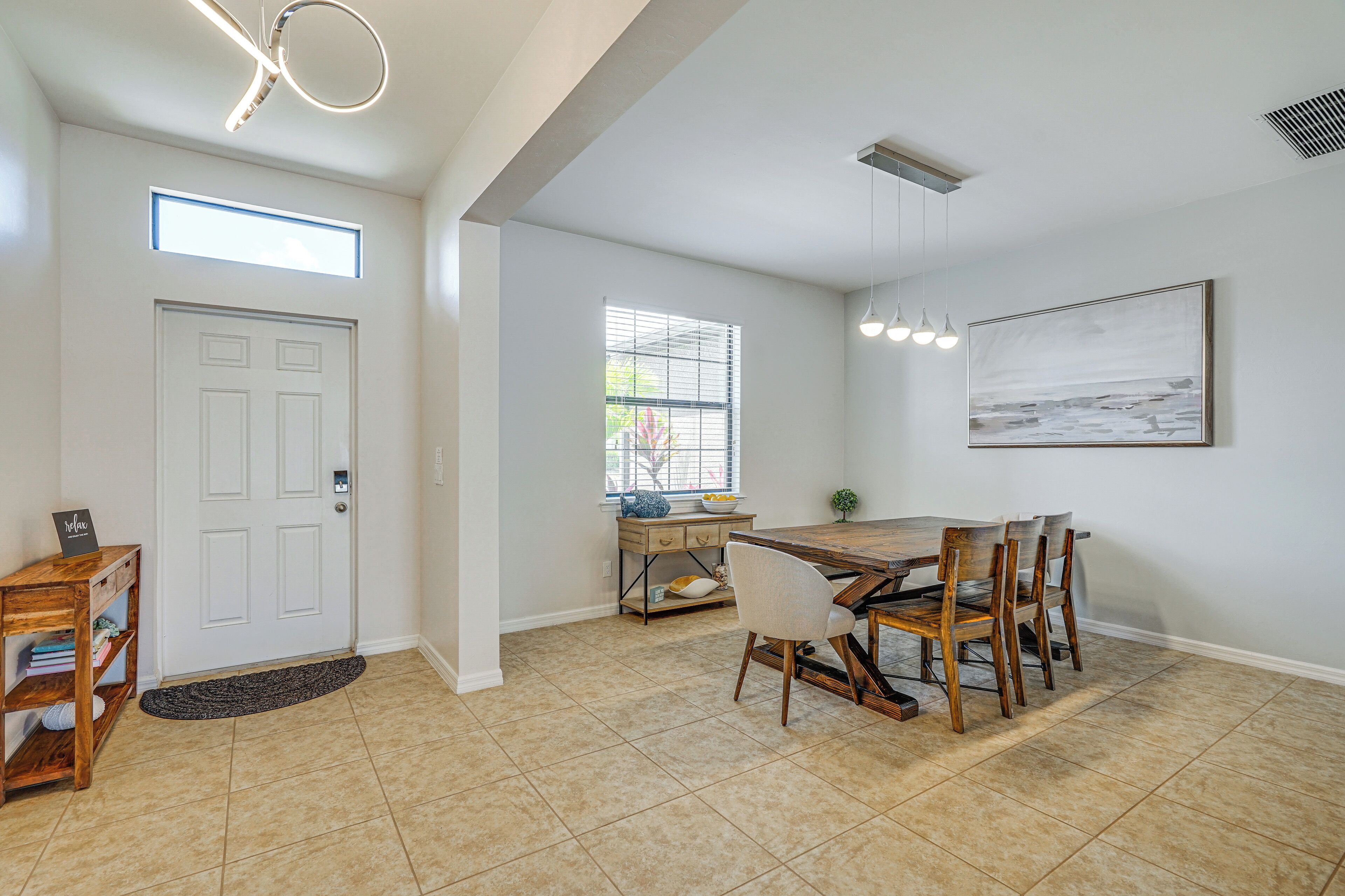 A spacious dining area with a wooden table and chairs, a small side table with a bowl and decorative items, a window with blinds, and a framed picture on the wall.