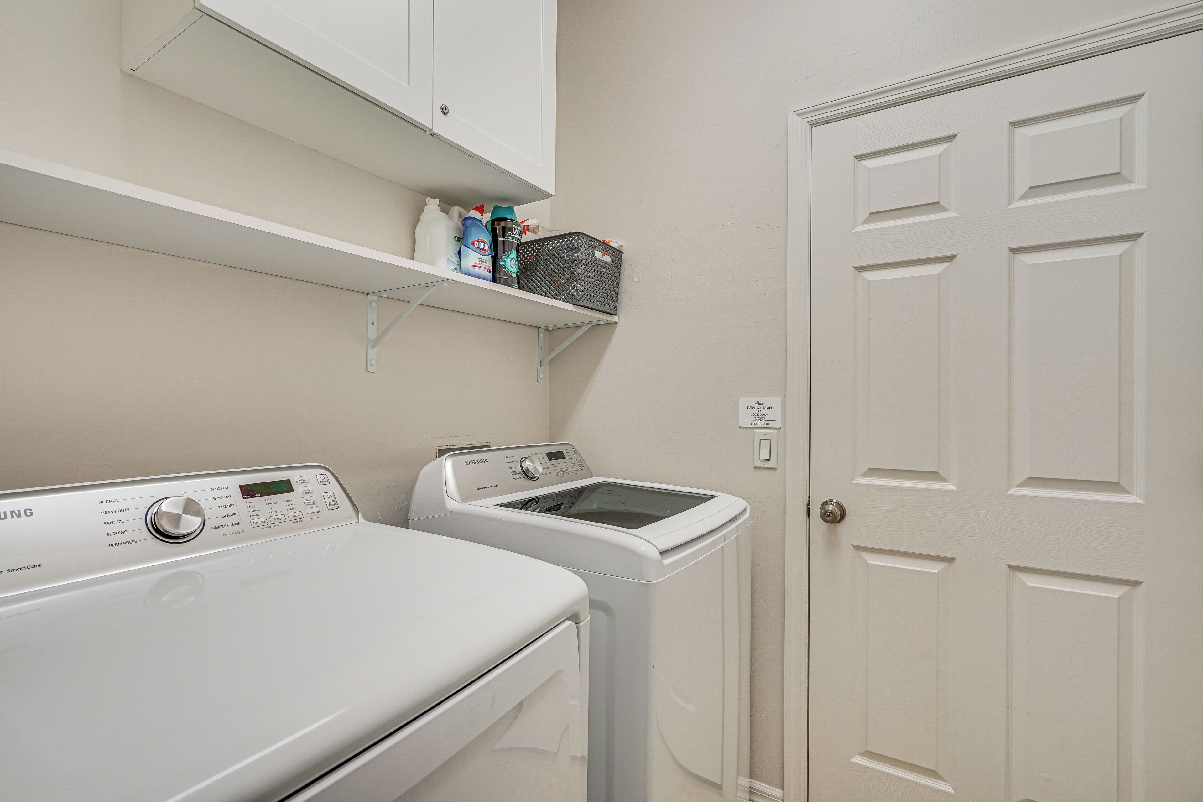 A laundry area with a front-loading washer and dryer, a shelf above the dryer with cleaning supplies, and a white door with a lock.