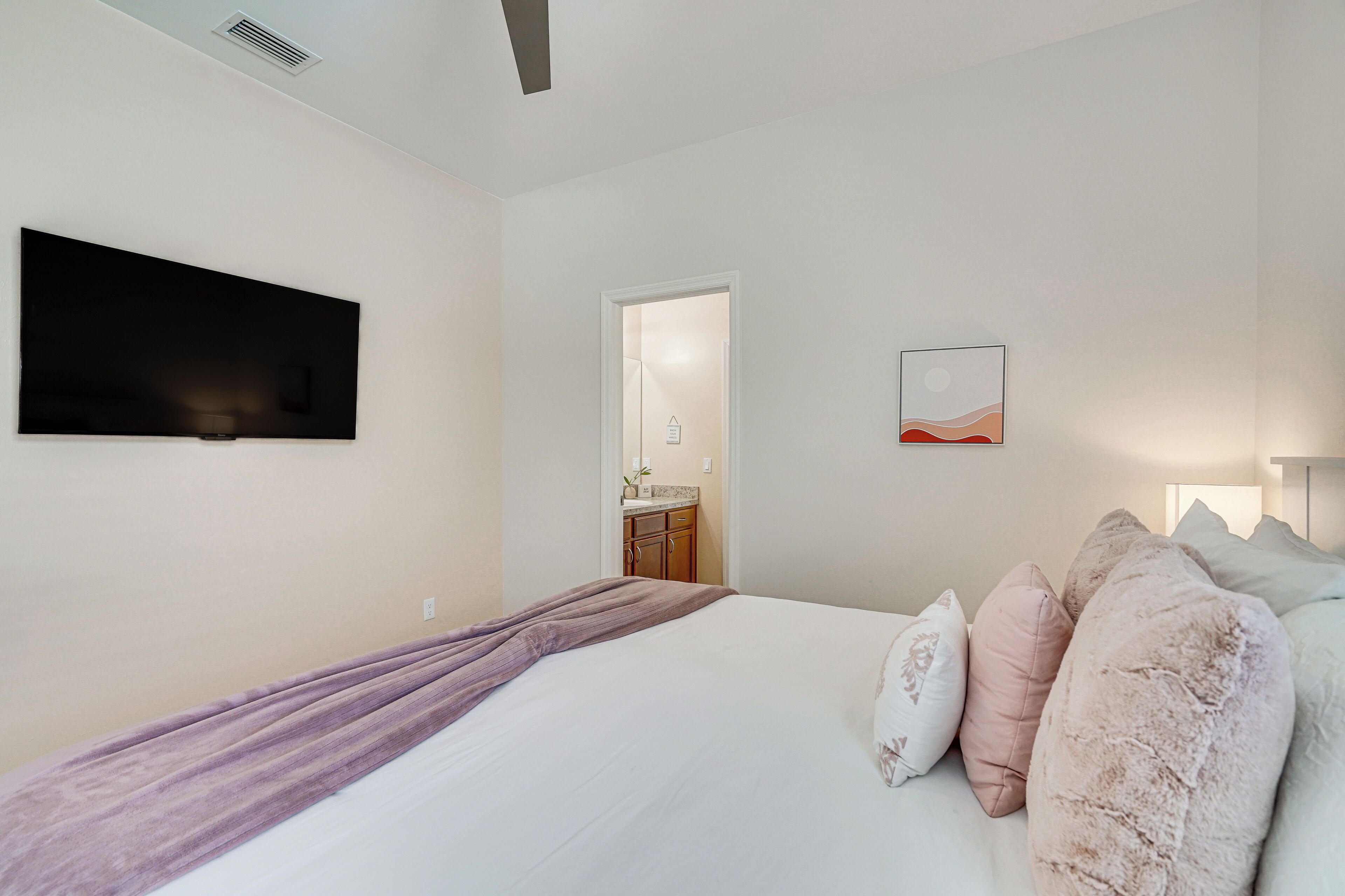 Un dormitorio con una cama, un televisor de pantalla plana montado en la pared, una puerta que da a un baño y una pequeña obra de arte en la pared.