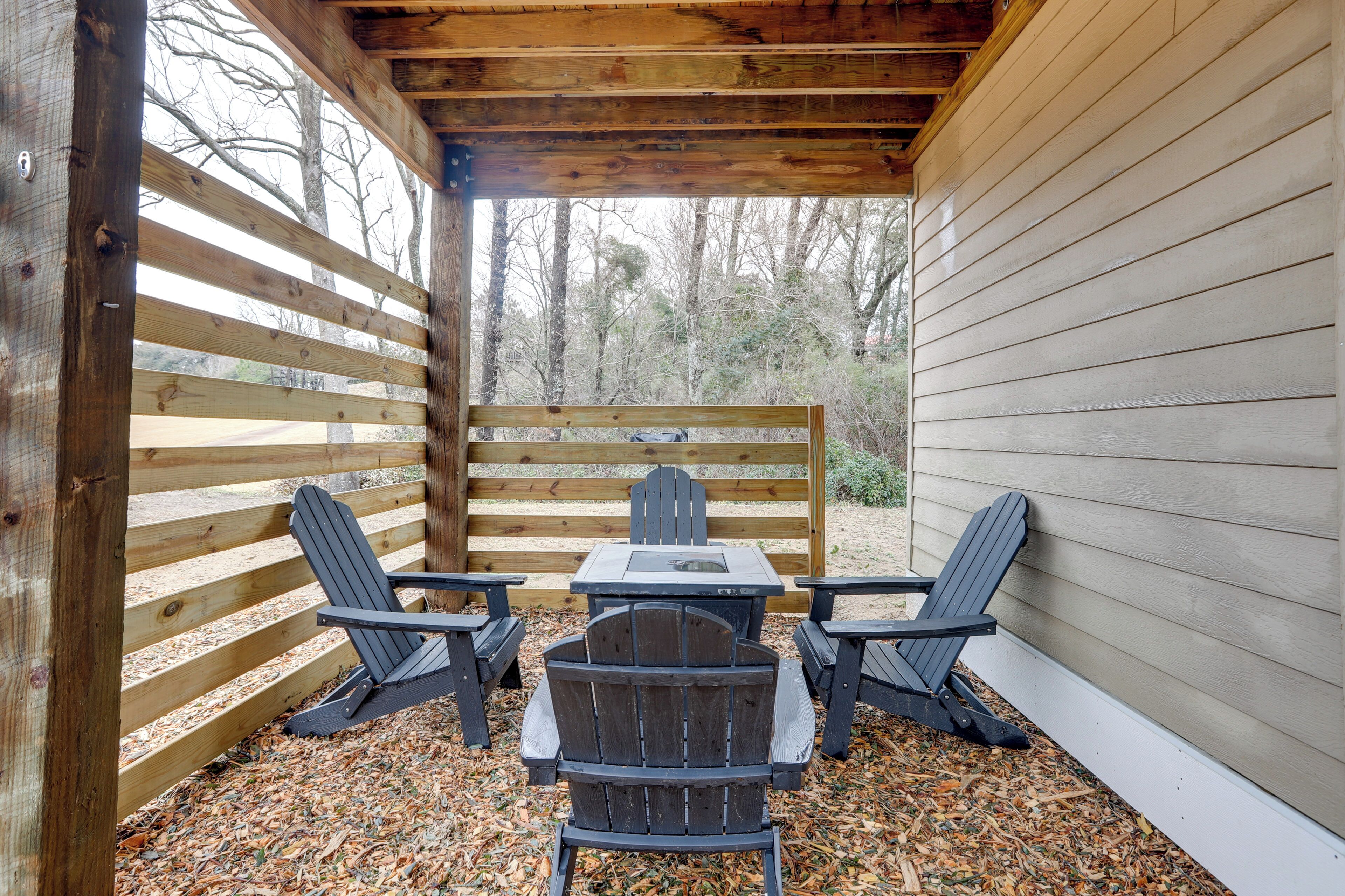 An outdoor patio with a wooden table and two Adirondack chairs, surrounded by a wooden fence and a white exterior wall.