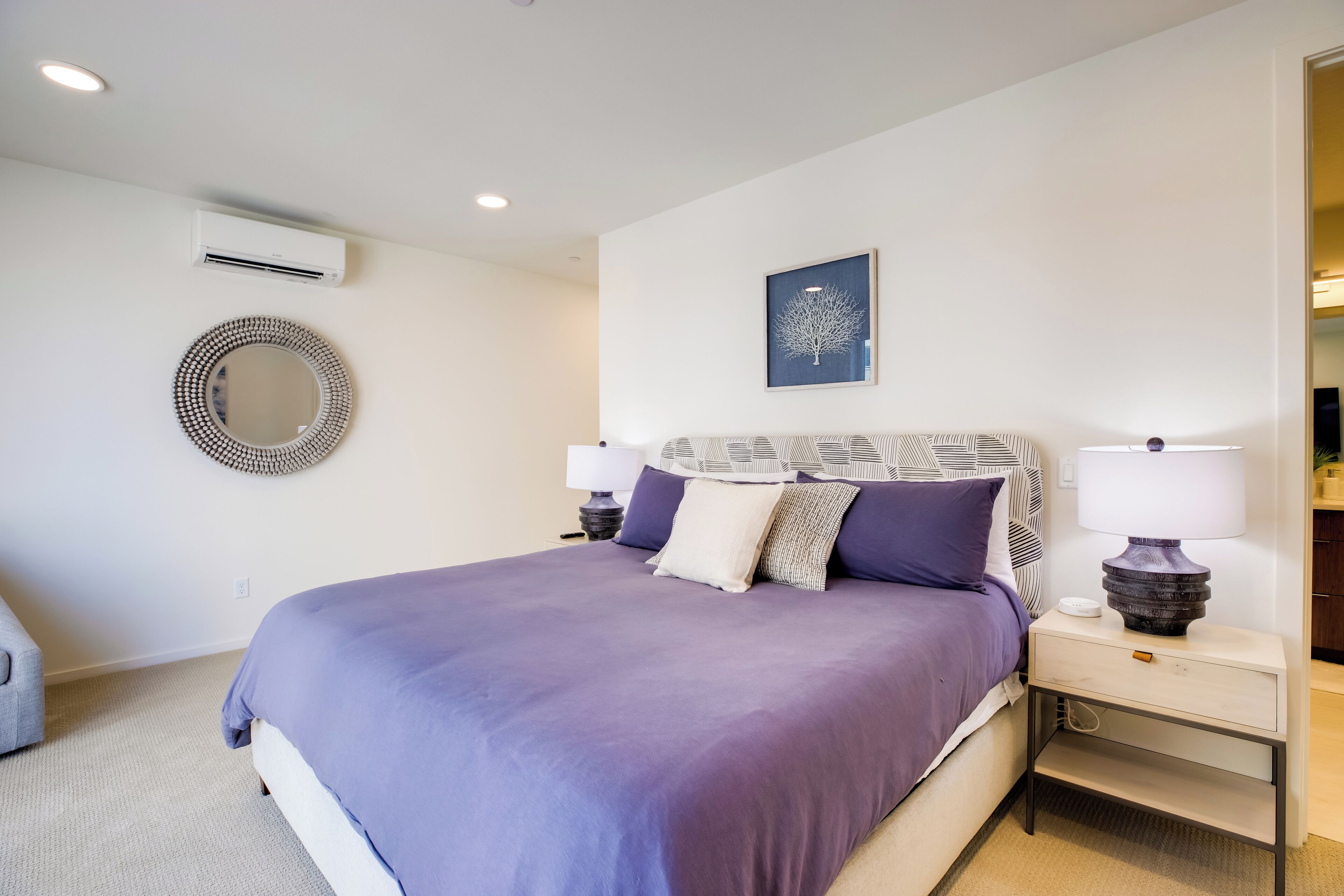 Un dormitorio moderno con una cama grande, dos mesitas de noche, un espejo redondo y un aire acondicionado de pared.