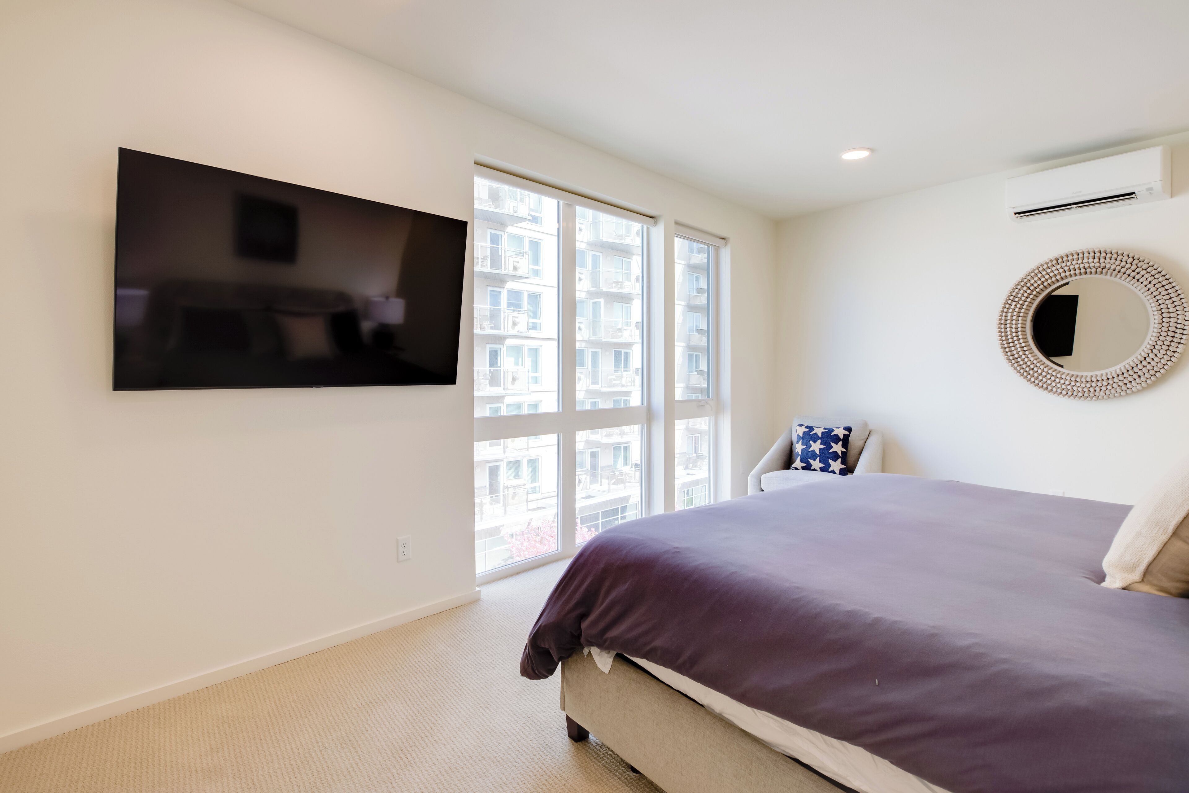 Ein Schlafzimmer mit einem großen Bett, einem flachbildfernseher an der Wand, einem Fenster mit Blick auf Gebäude und einem runden Spiegel.