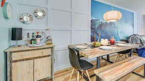 Un comedor con una mesa de madera, sillas negras y un banco. Hay relojes en la pared y un cuadro grande de una ola.