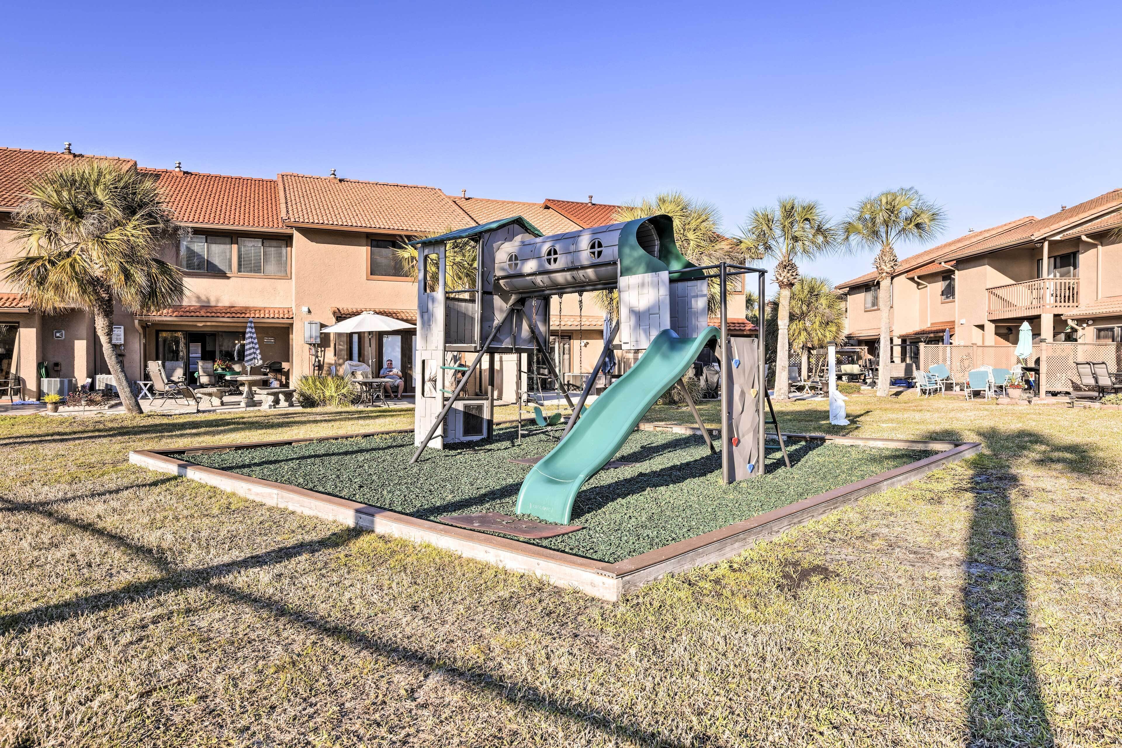 Un parque infantil con tobogán y estructuras de escalada, rodeado de césped y palmeras, con edificios al fondo.